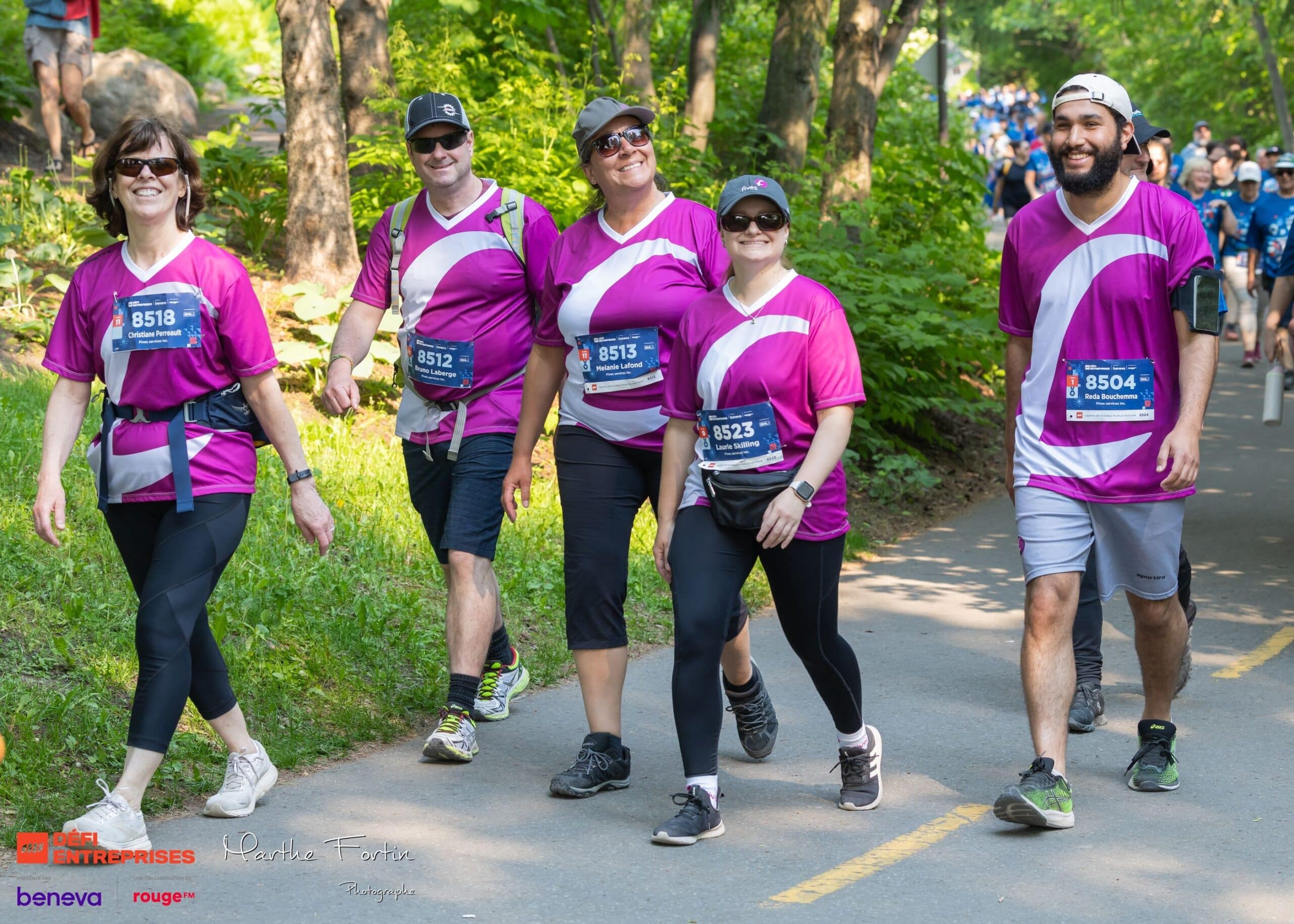 cmbrvpnu1003512pr5xa02sjd-processed | Défis Group of five runners in matching pink shirts walking on a tree-lined path during a race, bib numbers visible on chests.