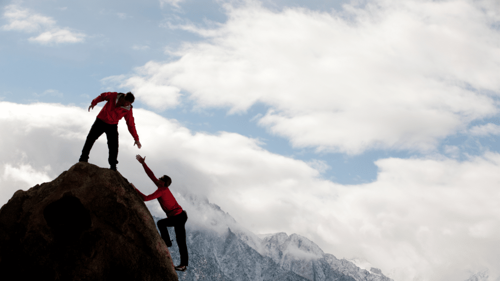Two climbers in red jackets reach each other on a rocky mountain summit against a cloudy blue sky.