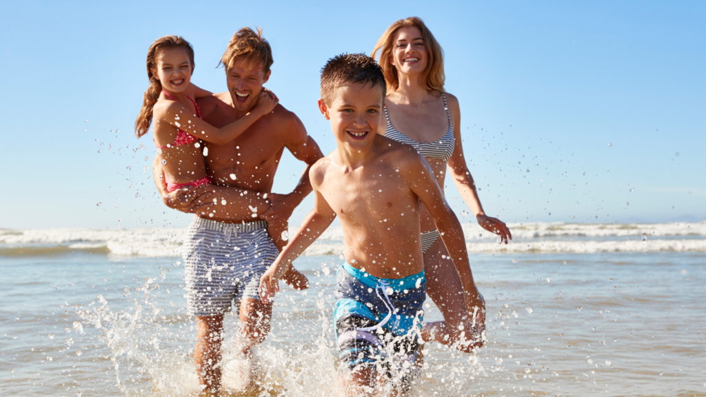 LEntraînement envacances - guide pratique 2 | Défis A family of four at the beach, two adults and two kids, splash in knee-deep water and smile at the camera.