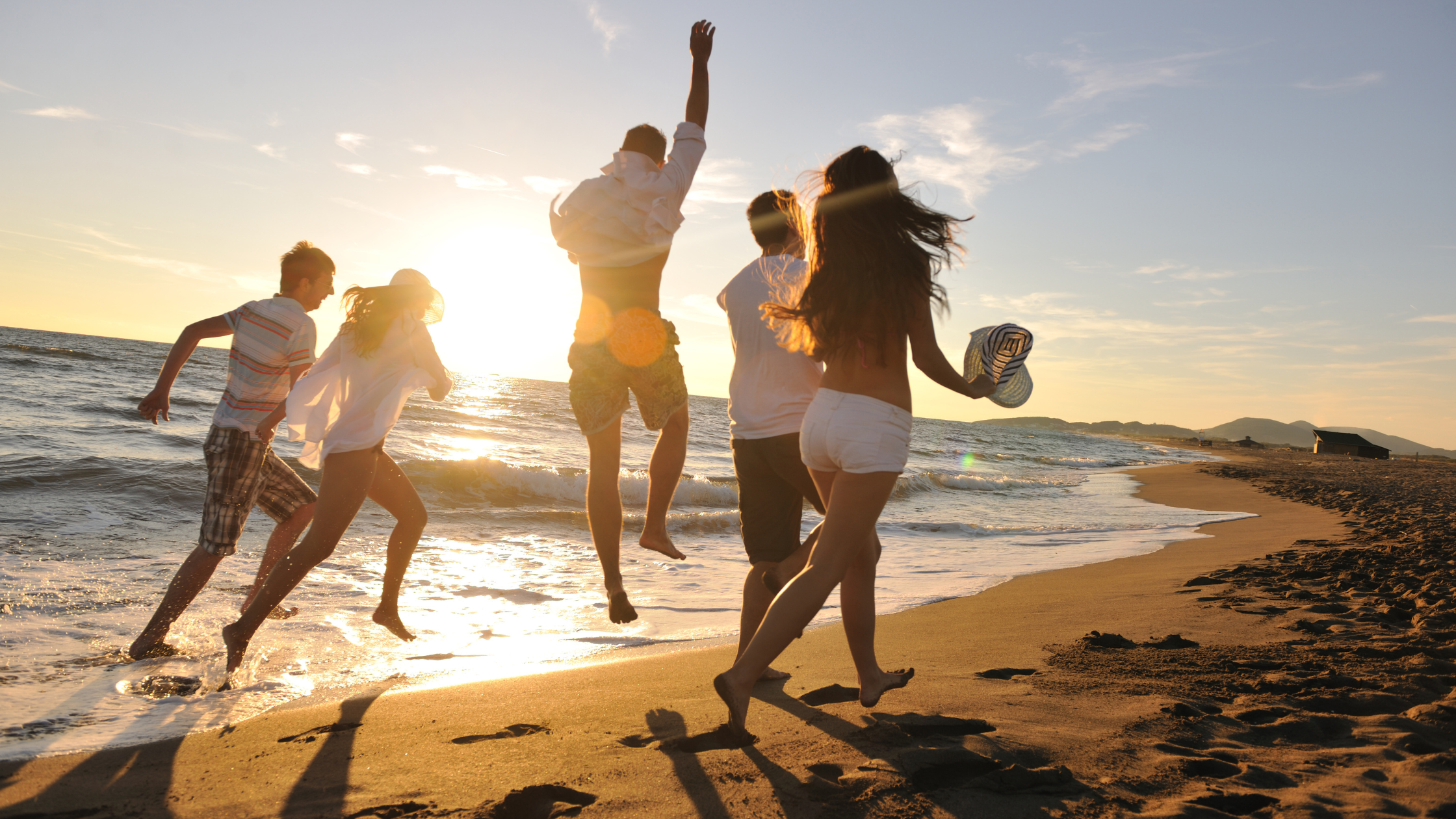 Group of friends jumping along a sunlit beach at sunset, waves behind them and footprints in the sand.