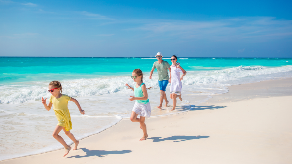 Lentraînement en vacances - guide pratique 3 | Défis Family of four running along a white sandy beach with turquoise ocean behind them under a clear blue sky, enjoying a sunny day (two adults and two children).