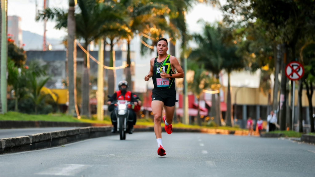 Male runner in neon-green and black singlet with bib 1182, sprinting toward the camera on a palm-tree-lined city street; motorcycle behind him.