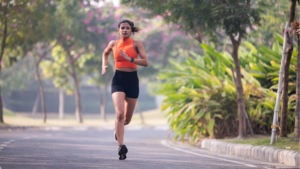 Woman running on a park path in an orange sleeveless top and black shorts, with greenery in the background.