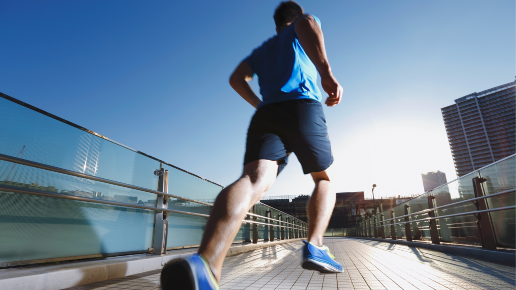 Runner viewed from ground level, wearing blue shirt and shorts, sprinting on a glass-floored urban pedestrian bridge under a clear blue sky.